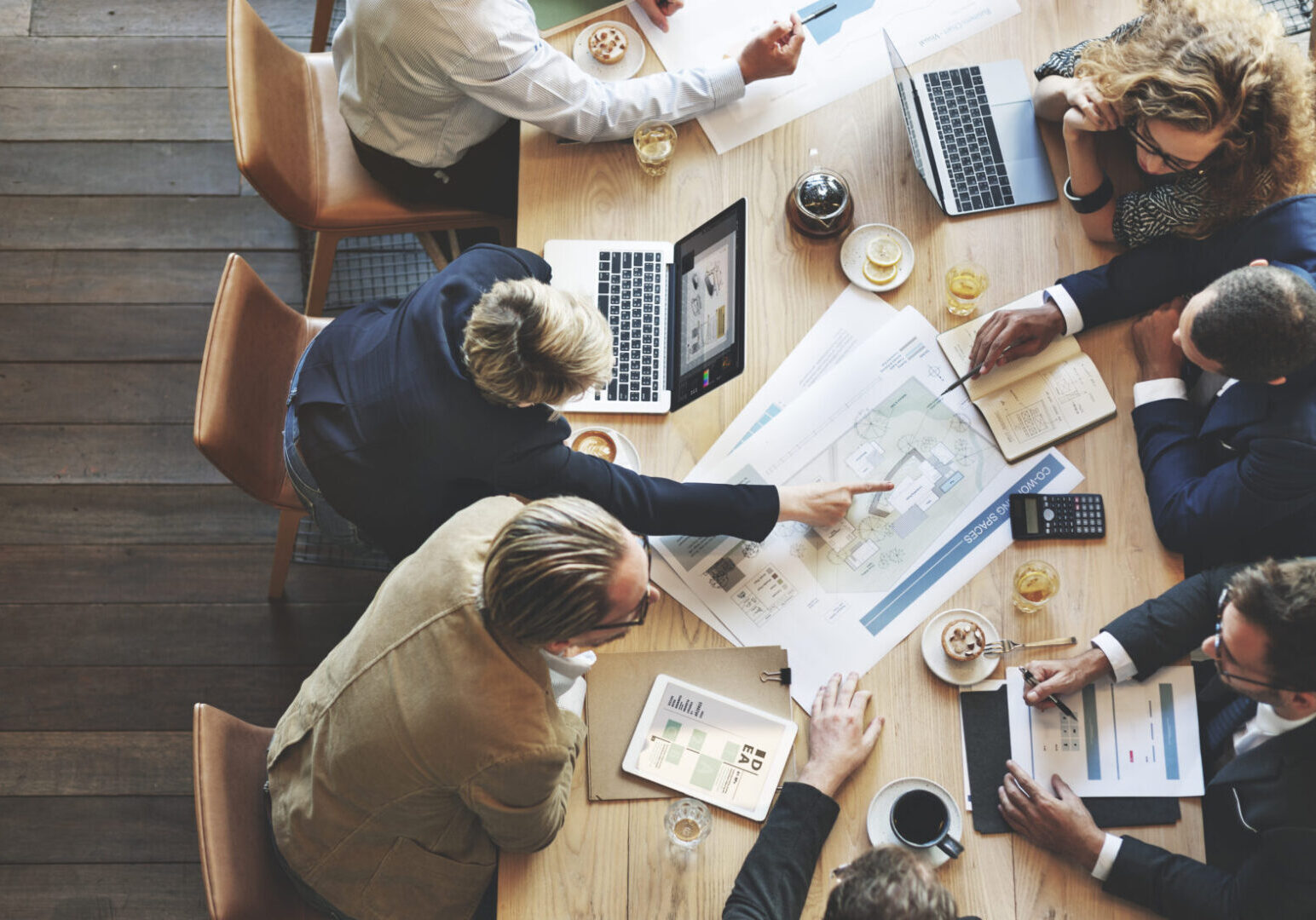 Six people sit around a wooden table with laptops, papers, and drinks, discussing documents and architectural plans in a collaborative meeting.